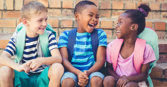 Three young children sitting together, wearing backpacks
