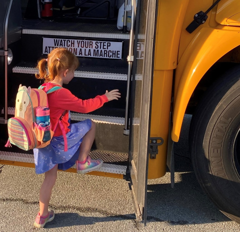 Young child with pigtails wearing a pink sweater, purple dress and backpack steps onto the school bus