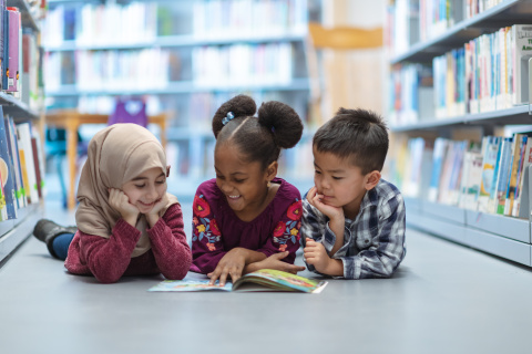 Three diverse elementary students laying on the floor of a library reading a book