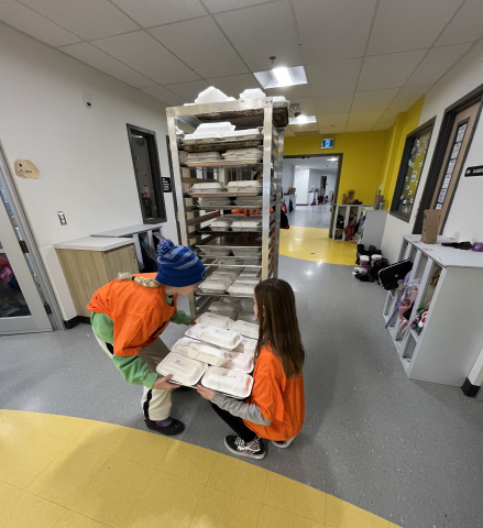 Photo of two student ambassadors in orange shirts pulling tray with lunches off cart in hallway of West Bedford School.