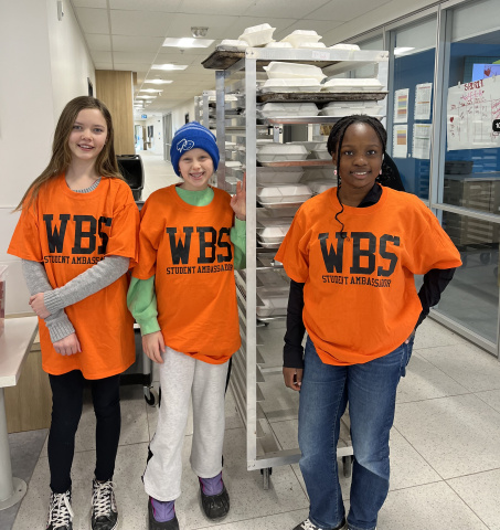 Photo shows three student ambassadors in orange shirts standing in front of a lunch cart at West Bedford School.